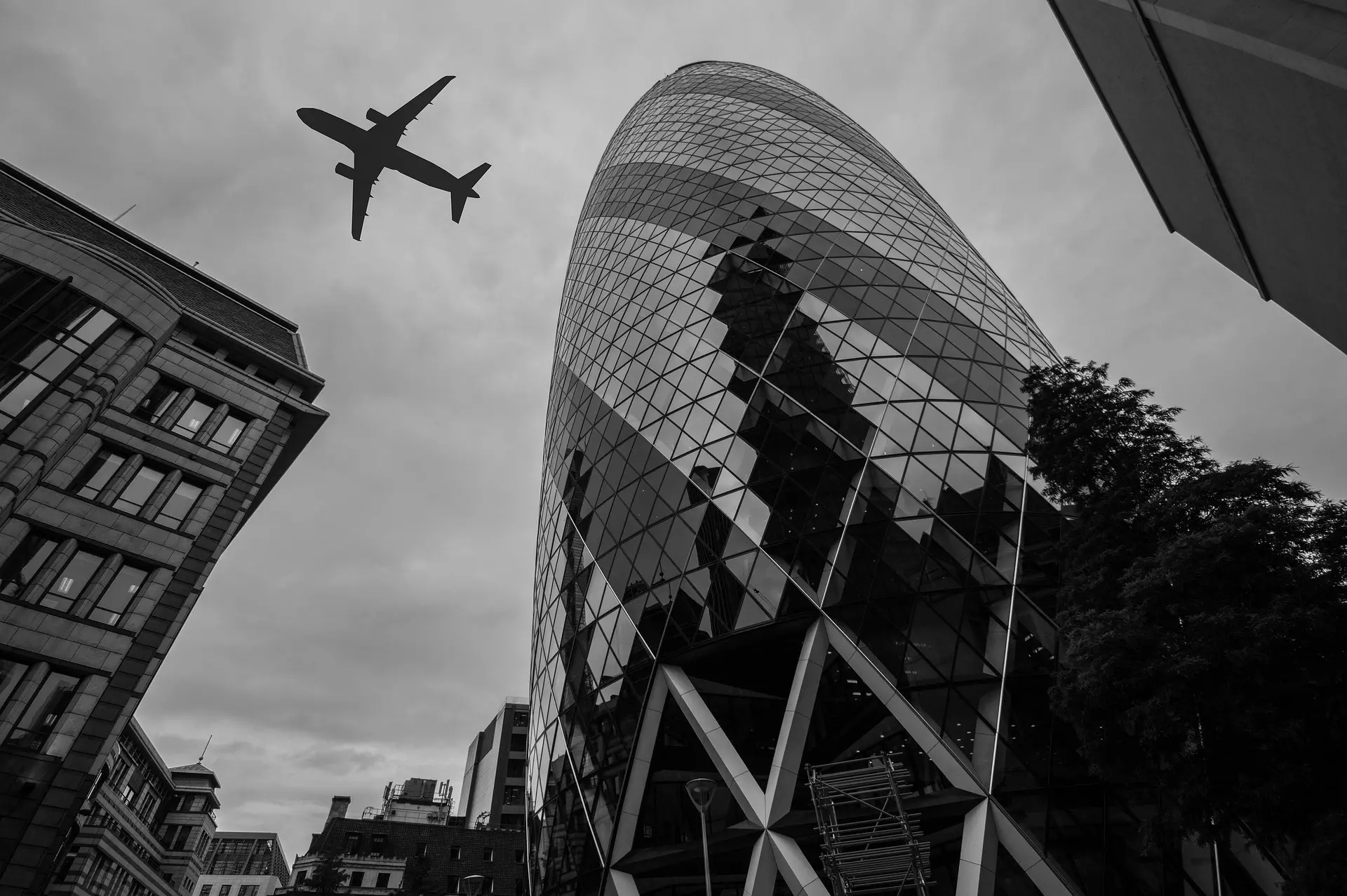 The Gherkin skyscraper viewed from below with an aircraft silhouetted against grey skies