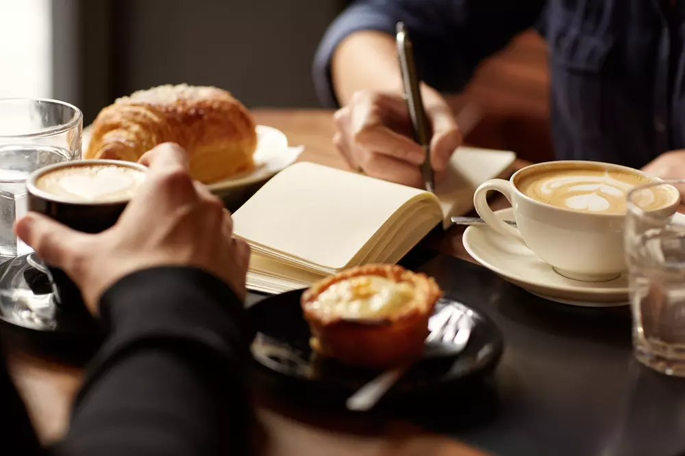 A person writes in a notebook over cups of coffee
